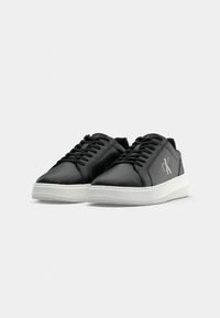 Zwarte synthetische sneakers met witte rubberen zolen, met tonale veters en een zilveren logodetail aan de zijkant. Overal een gladde textuur.
