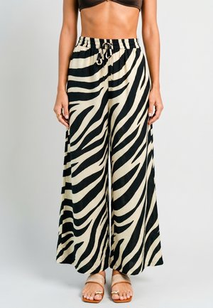 Vrouw draagt zwart-witte zebraprint wijde broek met trekkoord in de taille en beige sandalen met open tenen, staand tegen een effen achtergrond.