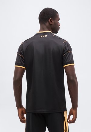 Mand iført sort sportsshirt med gule accenter og zigzag-mønstre på ærmerne, set bagfra mod en ensartet lys baggrund.