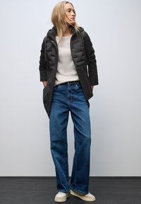 Black padded coat with hood layered over a beige sweater, paired with wide-leg blue jeans and light-colored sneakers. Smooth, casual look.