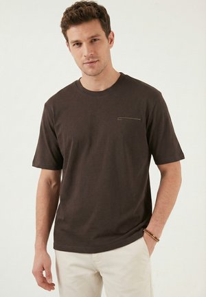 Młody mężczyzna o krótkich brązowych włosach, ubrany w ciemnobrązowy t-shirt z krótkim rękawem i jasnobeżowe spodnie, stojący na jednolitym tle.