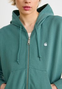 Teal zip-up hettegenser i mykt stoff med snor i hetta. Har lommer foran og en liten, hvit logo på brystet.