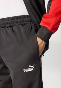 Sort træningsbukser med elastisk talje, prydet med et hvidt Puma-logo. En del af et tracksuit med en rød detalje på jakkeærmet.