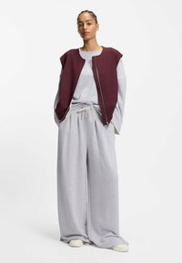 Grauer Baumwoll-Jogginganzug mit einer bordeauxfarbenen ärmellosen Jacke. Locker sitzende Hosen mit Falten, strukturierter Stoff und Kordelzugbund. Helle Schuhe.