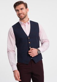 Gilet a quadri blu navy con accenti rossi, caratterizzato da scollatura a V, sei bottoni e due tasche laterali, indossato sopra una camicia rosa chiaro e pantaloni scuri.