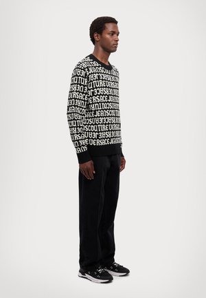 Maglione bianco e nero caratterizzato da un audace motivo testuale, colletto rotondo, polsini e orlo a costine. Abbinato a pantaloni scuri e sneakers nere.