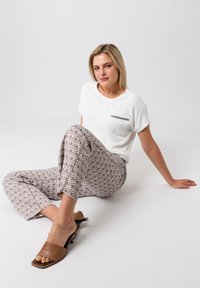 Un haut blanc à manches courtes avec une poche accent argentée, associé à un pantalon beige à motifs et des sandales à talons marron, assis sur un fond uni.