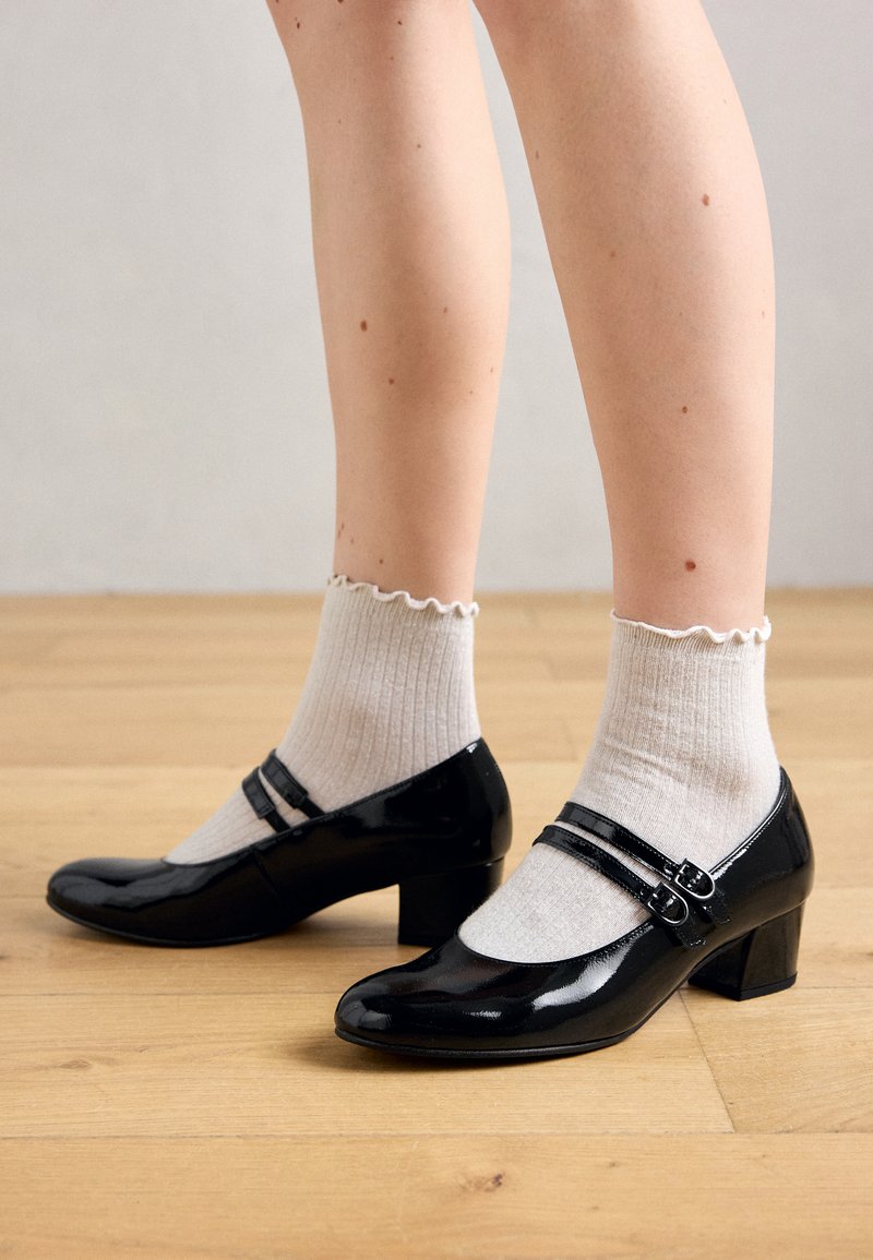 Jambes portant des chaussettes blanches côtelées avec des bords volants et des chaussures Mary Jane en cuir verni noir avec deux sangles à boucle, debout sur un sol en bois.