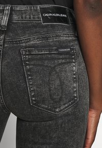 Jeans de mezclilla negra con un acabado desgastado. Presenta una etiqueta de Calvin Klein Jeans en la cintura y un diseño distintivo en el bolsillo trasero.