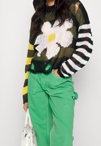 Pull en tricot avec un motif floral blanc et jaune, des rayures noires sur une manche, associé à un pantalon cargo vert et un sac blanc.