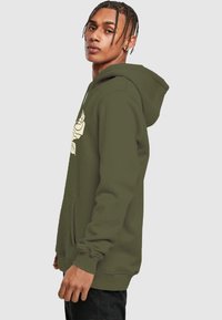 Olive spalvos džemperis su minkšta medžiaga, priekyje turintis kišenę, su dideliu baltu gėlių raštu krūtinėje ir su paminkštinta kapsule.