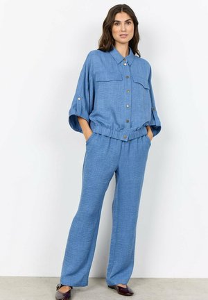 Vrouw staat met handen in de zakken, draagt een blauw overhemd met knopen en ellebooglange mouwen, gecombineerd met bijpassende wijde broekspijpen.