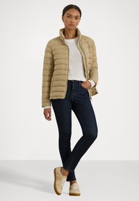 Giubbotto trapuntato beige con colletto alto, indossato sopra un maglione di lana bianco. Abbinato a jeans scuri e sneakers beige con lacci bianchi.