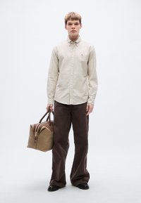 Πουκάμισο με κουμπιά σε κρεμ βαμβάκι, καφέ σκούρο παντελόνι με φαρδιά πόδια και δερμάτινη τσάντα duffel σε καφέ ταμπά με διπλές λαβές. Απλός και δομημένος σχεδιασμός.