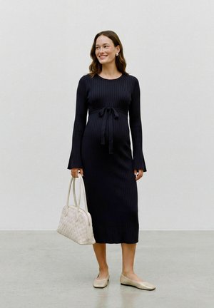 Pregnant woman smiling, wearing a long black ribbed dress tied at the waist, holding a white woven handbag and wearing light flat shoes.