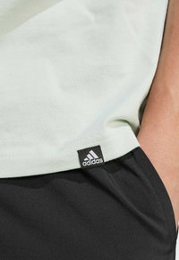 T-shirt in cotone verde chiaro con un'etichetta nera del logo Adidas, abbinato a pantaloni corti sportivi neri, che mostrano una texture liscia e un design semplice.