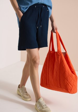 Person iført navy snøreløbeshorts, beige sneakers, der holder en stor quiltet knaldrød taske i hånden.