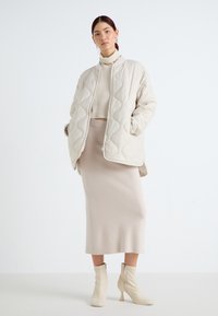 Quiltet beige jakke med høy krage, kombinert med en tettsittende beige genser og et matchende blyantskjørt, komplementert av kremfargede ankelstøvler.