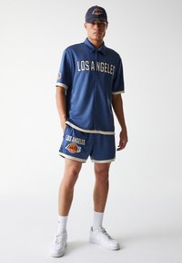 Lakers-themed outfit featuring a blue short-sleeve button-up jersey and matching shorts with white accents and logo, complemented by a dark cap.