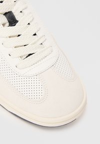 Baskets blanches avec un design perforé, tige en daim texturé, lacets plats et une semelle en caoutchouc solide. Esthétique minimaliste avec des lignes épurées.
