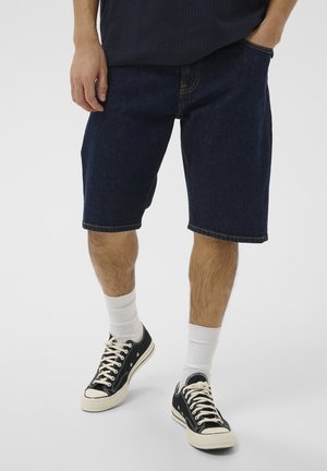 Persoon die donkerblauwe spijkershorts draagt, witte enkelsokken en zwarte lage canvas sneakers met beige zolen en veters.