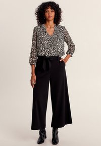 Schwarz-weiß gemusterte Bluse mit Dreiviertelärmeln, kombiniert mit schwarzen weit geschnittenen Hosen und schwarzen Ankle Boots.