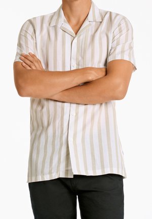 Personne les bras croisés portant une chemise beige et blanche à manches courtes à rayures verticales et un pantalon noir.