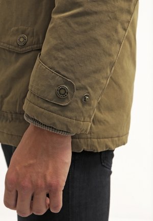 Ärmel einer braunen Barbour-Jacke mit geknöpftem Riemen, Bündchen eines grauen Strickpullovers sichtbar, der auf einer schwarzen Hose ruht.