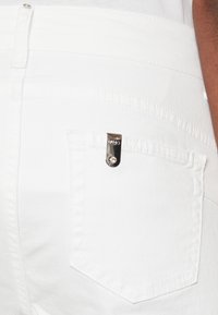 Jeans in denim bianco con una tasca a toppa che presenta un dettaglio in metallo argentato. Texture liscia con cuciture visibili lungo le cuciture.