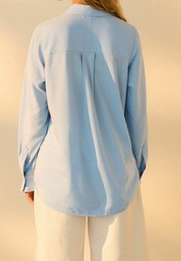 Camicia azzurro chiaro a maniche lunghe con colletto, dettaglio della piega posteriore e polsini abbottonati, abbinata a pantaloni bianchi in tessuto morbido.