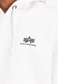 White hoodie made from soft fabric featuring a black logo with angular lines and text "ALPHA INDUSTRIES" near the chest area.