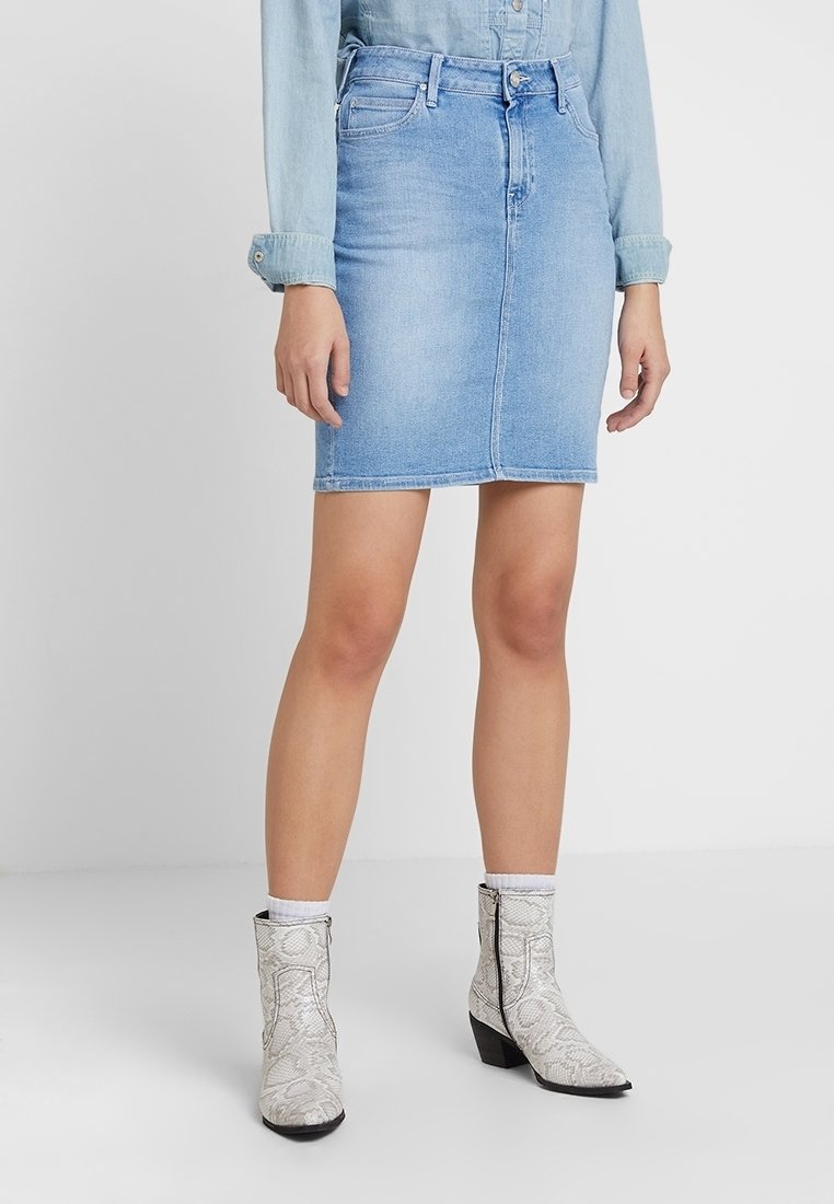 Ljusblå denimkjol med rak skärning, mitt längd, med bältesöglor, framknäppning och subtil blekning. Kombinerad med vita mönstrade stövlar.