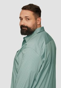 Koszula z krótkim rękawem w jasnym turkusie z geometrycznym wzorem. Posiada kołnierzyk zapinany na guziki oraz luźny krój. Tkanina wydaje się gładka.