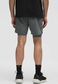 Une personne se tient de dos, portant un t-shirt noir, un short de sport gris et des chaussettes noires avec des baskets, sur un fond uni.