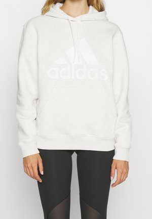 Femme portant un sweat à capuche blanc Adidas avec logo sur le devant et un legging noir avec des panneaux transparents sur les cuisses.