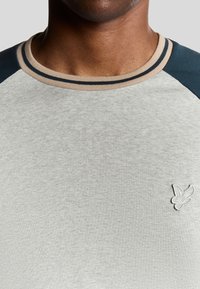 Lys grå t-shirt med korte ærmer, marineblå skulderakcenter, ribbet krave med beige og mørkeblå striber, der har et lille broderet logo.