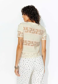 Femme aux cheveux bouclés et foncés, portant un haut en dentelle crème et un pantalon blanc avec de petits motifs floraux bleus, se tenant devant un fond blanc.