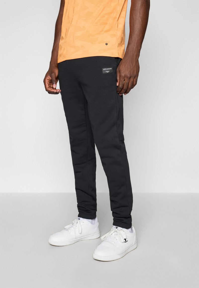 Calças de moletom pretas com um corte justo, apresentando um pequeno logótipo. Combinadas com uma t-shirt laranja estampada e ténis brancos.