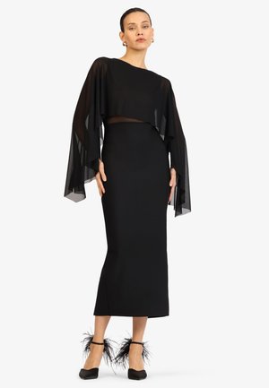 Frau steht, trägt ein langes schwarzes Kleid mit durchscheinenden, drapierten Ärmeln und schwarze Absatzschuhe, verziert mit schwarzen Federakzenten.