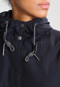 Žena na sobě má tmavě modrou bundu s kapucí, kterou zdobí šedé stahovací šňůrky svázané do uzlů a kovové knoflíky s nápisem "Lee Since 1889".