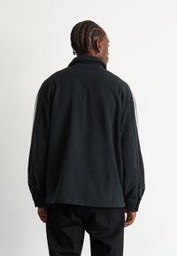 Černý fleecový pulovr s vysokým límcem, zdobený dvěma bílými pruhy podél rukávů a uvolněným, oversized střihem.