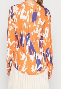 Košile s dlouhým rukávem a uvolněným střihem, s abstraktním vzorem v oranžové, bílé a fialové barvě, vyrobená z lehkého materiálu, s kulatým lemem.