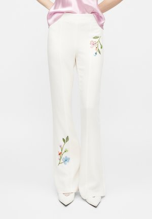 Λευκό παντελόνι με φαρδύ πόδι και πολύχρωμη κεντητή λουλουδάτη διακόσμηση στο πάνω μέρος του μηρού και στο κάτω μέρος του ποδιού, φορεμένο με ροζ μπλούζα και λευκά μυτερά παπούτσια.