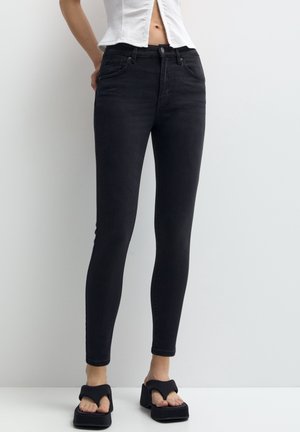 Jean noir slim porté avec des sandales à plateforme noires et un haut court blanc à boutons, debout devant un fond gris clair uni.