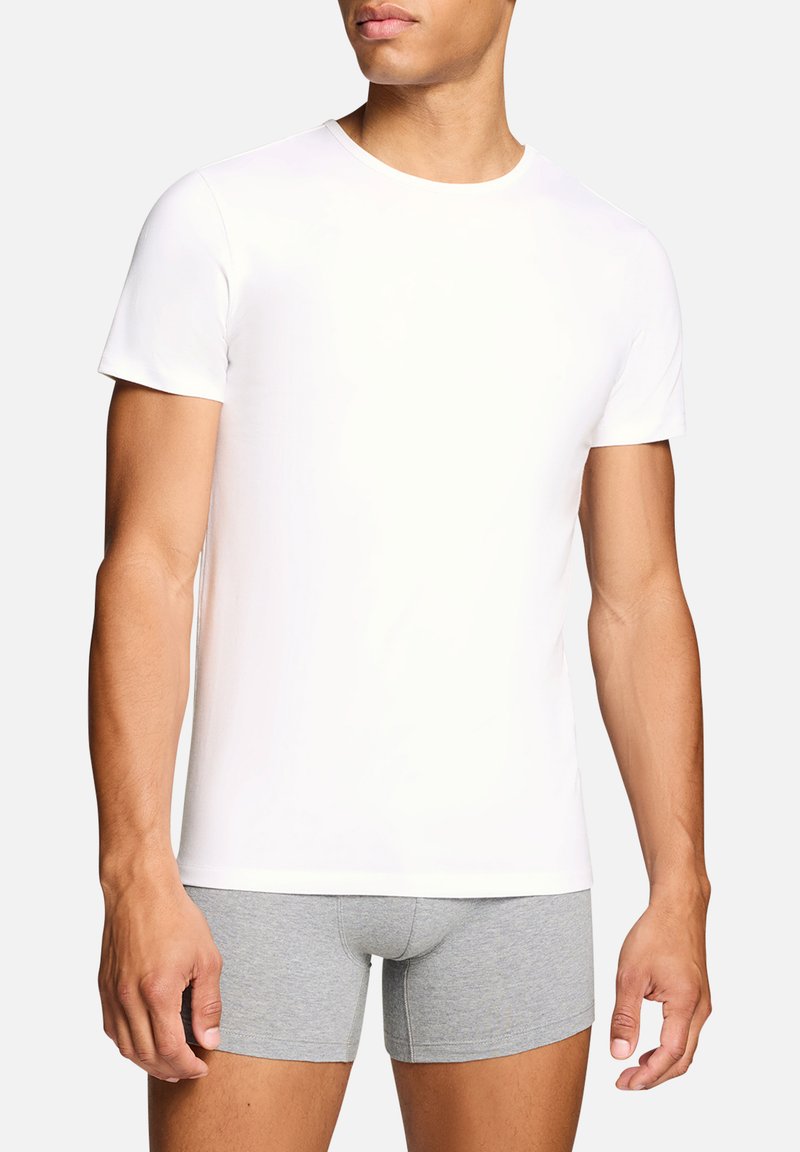 Jeune homme portant un t-shirt blanc uni à manches courtes et un boxer gris clair, debout devant un fond uni.