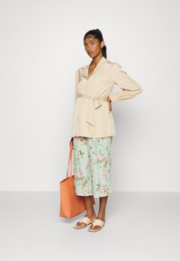 Camisa beige de botões com cintura amarrada, calças largas florais em menta e uma tote laranja. Sandálias leves completam o look. Texturas suaves presentes.