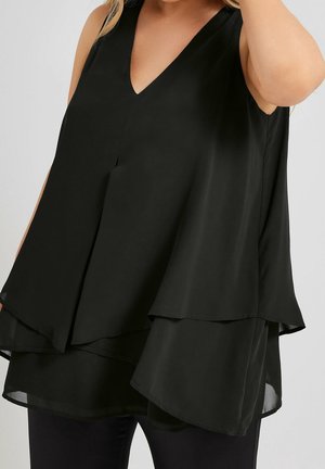 Femme portant un haut noir sans manches à couches superposées avec un col en V et une coupe ample, associée à un pantalon noir sur un fond uni.
