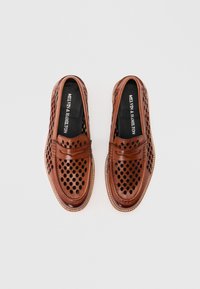 Καφέ δερμάτινα μοκασίνια με διάτρητες οπές και μαύρα πέλματα, τοποθετημένα δίπλα δίπλα σε λευκό φόντο.