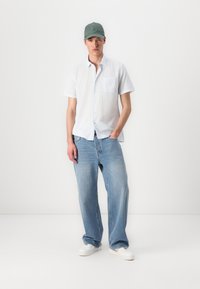 Camisa branca de manga curta com bolso no peito, combinada com jeans de corte largo de tom azul claro e ténis brancos. Um boné verde-azeitona completa o visual.