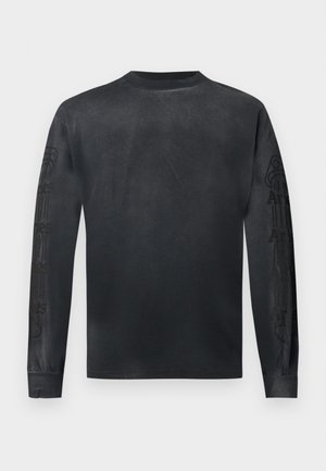 Young man standing wearing black long-sleeve shirt, faded blue jeans with light graphic prints, and brown leather boots with orange laces.